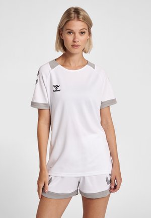 Blondynka w białym stroju sportowym z szarymi lamówkami, stojąca na jednolitym jasnoszarym tle, zwrócona przodem.