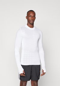 Langärmliges Sportshirt in Weiß mit einem strukturierten Gittermuster und verlängerten Ärmelbündchen, kombiniert mit schwarzen Shorts für den Kontrast.