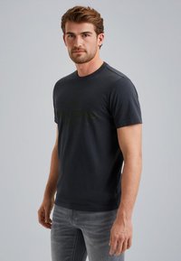 Camiseta de algodón negra con mangas cortas, escote redondo, que presenta un sutil diseño de logo verde en la parte delantera. Combinada con jeans de color gris claro.