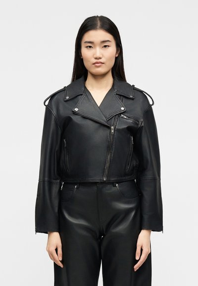 Femme aux cheveux bruns et raides portant une veste en cuir noire courte avec des fermetures éclair argentées et un pantalon en cuir noir, debout devant un fond uni.
