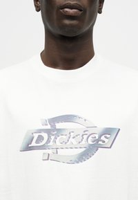 Κοντινό πλάνο ενός ατόμου που φοράει λευκό πουκάμισο με ένα λαμπερό, μεταλλικό λογότυπο «Dickies» τυπωμένο στο μπροστινό μέρος.