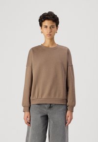 Sudadera marrón hecha de tejido suave, con cuello redondo, mangas largas y dobladillo acanalado. Llevada con unos vaqueros grises holgados.