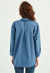 Camicia blu a maniche lunghe con colletto, dettaglio con piega sul retro e polsini abbottonati. Tessuto dalla superficie liscia. Pantaloni parzialmente visibili sotto.