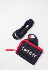 Chaussons pliables noirs Tommy avec motif d'étoiles rouges, l'un à l'intérieur d'une pochette assortie avec fermeture éclair rouge et logo de marque blanc et rouge.
