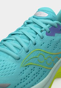 Κοντινό πλάνο τουρκουάζ αθλητικού παπουτσιού με κίτρινο λογότυπο, μοβ λεπτομέρεια οπής, διαπνέον δίχτυ και κορδόνια στο ίδιο τουρκουάζ χρώμα.