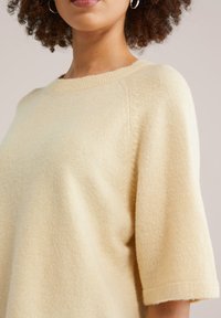 Femme aux cheveux bouclés portant un pull en maille crème à manches courtes et des boucles d'oreilles argentées en forme de créoles sur un fond uni.