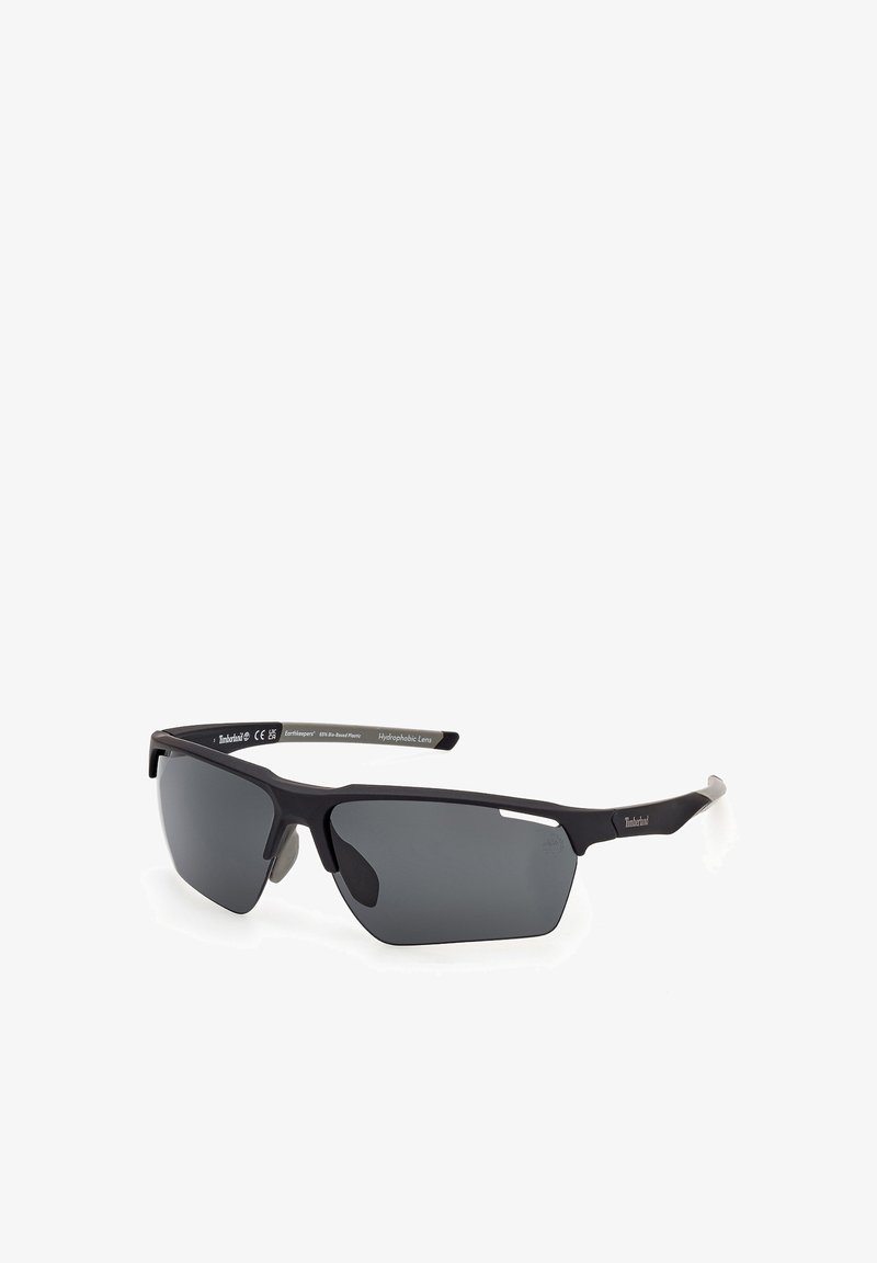 Lunettes de soleil semi-cerclées noires avec des verres rectangulaires foncés et des branches fines, arborant le logo Timberland sur la branche, présentées sur un fond blanc.