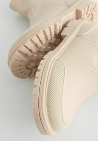 Bottes ankle beiges en matériau synthétique avec des semelles texturées. Présente un dessus lisse et une semelle en caoutchouc à motifs.