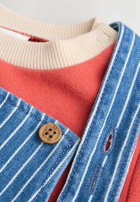Stripede blå arbejdstøj med en træknap, lagdelt over en koralfarvet sweater. Ribbet beige halsudskæring, der har strukturerede stoffer og synlige sømme.