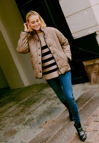 Vadderad beige jacka med tryckknappar, bärs över en svart-och-beige randig tröja, kombinerad med blå jeans och svarta ankle boots.