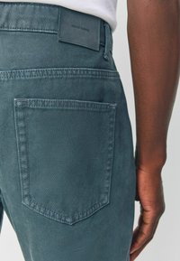 Džínové kalhoty v tmavě modrozelené barvě. Texturovaná tkanina s zadní kapsou a koženým štítkem s nápisem "DENIM SUPPLY" na pase.