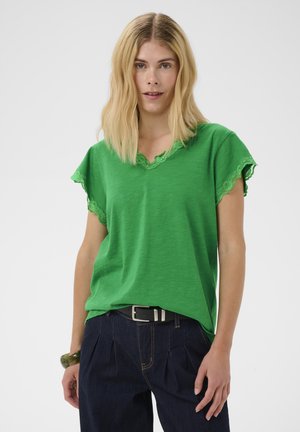 Femme blonde portant un haut vert à manches courtes avec bordure en dentelle et un pantalon plissé bleu foncé avec une ceinture noire, debout sur fond blanc.