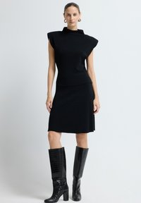 Schwarzes Strickkleid mit einem hohen Kragen und strukturierten Schultern, kombiniert mit glänzenden schwarzen kniehohen Stiefeln mit Krokodilprägung.