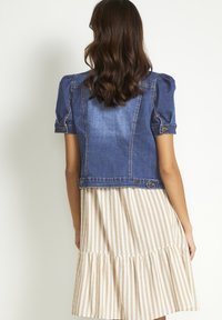 Jeansjacka med kort puffärmar, blå färg och avslappnad design. Stylad över en beige randig klänning med tierad nederkant.