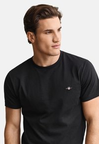 Junger Mann mit gestylten braunen Haaren, der ein schwarzes GANT-T-Shirt trägt und leicht nach rechts blickt vor hellem, einfarbigem Hintergrund.