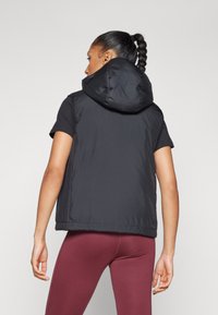 Svart pufferväst med stor huva, quiltad struktur, utan ärmar och justerbar nederkant, tillsammans med åtsittande bordeauxfärgade leggings.