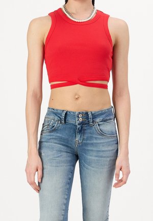 Femme portant un crop top rouge sans manches avec détail drapé et collier à plusieurs rangs, associé à un jean bleu délavé taille haute.