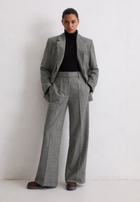 Costume gris sur mesure avec pantalon à jambes larges et col roulé noir ajusté. Le matériau présente un motif texturé ; la veste a des revers à crans.