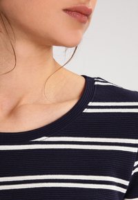 Top de punto a rayas azul marino con rayas horizontales blancas, escote redondo y textura acanalada. Vista en primer plano del área del hombro y el cuello.