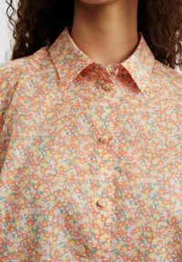 Camicia a maniche corte con bottoni, presenta un motivo floreale in tonalità arancione pastello, giallo e verde, con colletto e tessuto morbido e strutturato.
