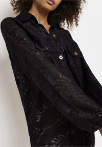 Camicetta nera a maniche lunghe con texture e motivo in pizzo, dotata di due tasche frontali e bottoni metallici, che mostra una leggera lucentezza.