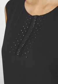 Haut sans manches noir avec un décolleté en trou de serrure, ornée d'accents métalliques et de petits clous disposés en motif autour de la patte de boutonnage.