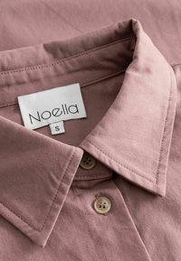 Putns rozā krekls ar strukturētu apkakli, divām pogām un gludu tekstūru. Uz balta birkas redzama uzraksts "Noella".