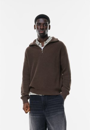 Brun ribbet striktrøje med halv-zip krave, lagd over en lys ternet skjorte, parret med mørke denim jeans.