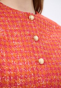 Tessuto testurizzato in tonalità di rosa e arancione con tre bottoni rotondi dorati e scanalati. Primo piano di un design con colletto.