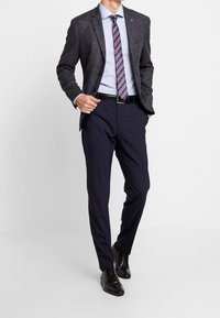 Ciemny, teksturowany żakiet z dopasowanym krojem na jasnoniebieskiej koszuli, w połączeniu z granatowymi spodniami i czarnymi eleganckimi butami. Pasek w paski dodaje kontrastu.