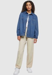 Donkerblauwe spijkerjas met knopenlijst, puntige kraag en twee borstzakken. Gepaarde met beige wijde broek en witte sneakers.