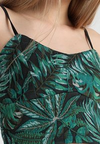 Haut tropical vert à motif de feuilles sur fond noir, avec fines bretelles et tissu texturé.