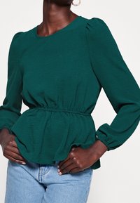 Blouse verte texturée à manches longues, taille élastique et détails froncés. Portée avec un jean délavé clair, mains sur les hanches.