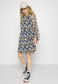 Vestido floral com fundo azul-marinho, apresentando flores amarelas e brancas, mangas longas e transparentes, cintura justa e comprimento acima do joelho. Combinado com ténis pretos.