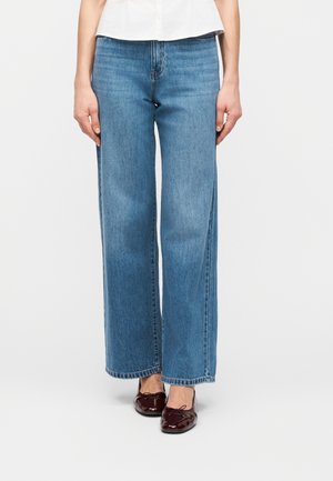 Kvinne iført høytlivs blå jeans med rette ben, hvit topp og skinnende mørkerøde ballettsko på hvit bakgrunn.