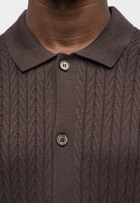 Σκούρο καφέ πλεκτό πουλόβερ πόλο με υφασμάτινο σχέδιο σε σχήμα ψαροκόκαλου και δύο ορατά κουμπιά, φορεμένο από άτομο με μέτριο σκούρο τόνο δέρματος.