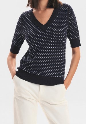 Vrouw draagt een zwart kort mouws V-hals trui met kleine lichtblauwe stippen, gecombineerd met witte hoge taille broek, handen in de zakken.