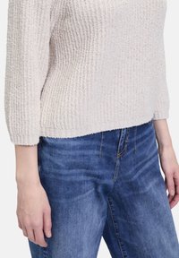 Light beige textured knit sweater with three-quarter sleeves, paired with blue denim jeans featuring a slight fade and visible stitching.