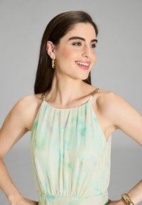 Hellgrünes, Batik-Halterkleid mit gerafften Details und goldenen Hardware-Elementen an den Trägern. Modell trägt goldene Creolen.