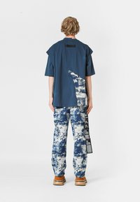 Persona con cabello rizado corto de pie, vistiendo una camisa de manga corta azul marino y pantalones de patrón blanco y azul con zapatos marrones.