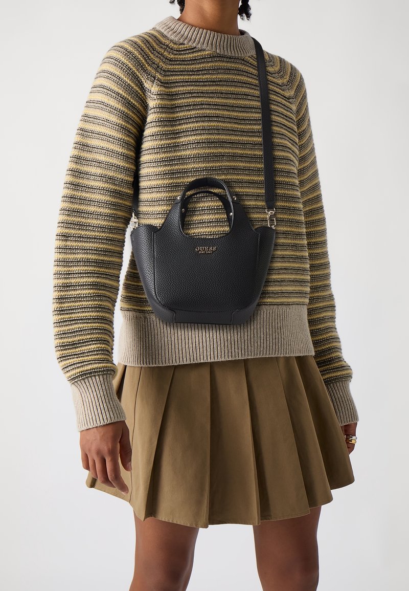 Personne portant un pull en maille rayé et une jupe plissée olive, portant un petit sac à bandoulière noir Guess en travers de la poitrine.