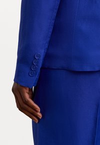 Manche d'un blazer en lin bleu avec cinq boutons assortis. Texture lisse avec une coupe ajustée. Gros plan sur une main reposant sur le vêtement.