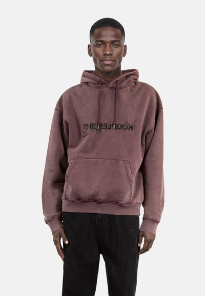 Bruine oversized hoodie met een voorzak, met een getextureerde zwarte afdruk van de woorden "THE KOOKIED" op de borst.