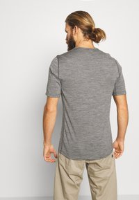 Άνδρας με μούσι και μαλλιά δεμένα πίσω, φοράει εφαρμοστό γκρι κοντομάνικο πουκάμισο και μπεζ παντελόνι, στέκεται αντικρίζοντας έναν λευκό τοίχο.