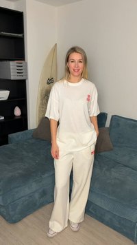 Γυναίκα με άσπρο υπερμεγέθες t-shirt και φαρδιά παντελόνια στέκεται μπροστά σε μπλε καναπέ με σανίδα του σερφ που στηρίζεται στον τοίχο κοντά της.