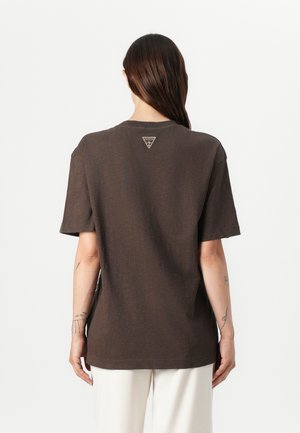 Persoon met lang donker haar draagt een losse donkerbruine T-shirt met een klein lichtgekleurd logo aan de bovenrug, gecombineerd met een witte broek.