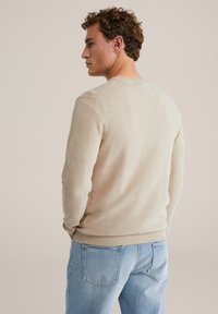 Bež teksturirani džemper s okruglim izrezom i rebrastim manšetama i rubom, uparen s svijetloplavim trapericama, pogled sa stražnje strane.