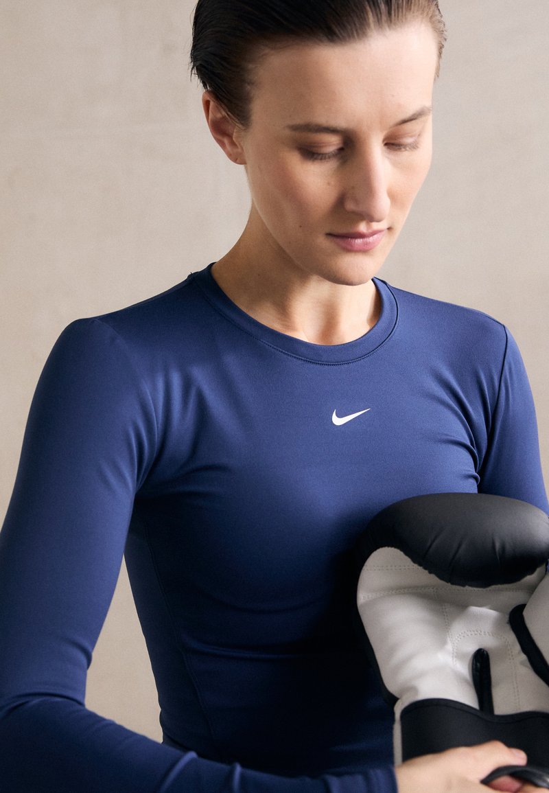 Tamno plavi dugi rukav sportska majica s okruglim izrezom i bijelim Nike logotipom, uparen s crnim i bijelim boksačkim rukavicama koje se drže u rukama.
