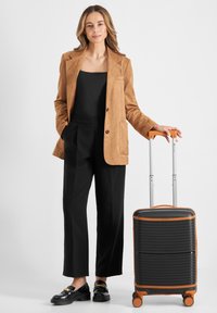 Blazer in suede marrone, topp black e pantaloni neri su misura. Valigia nera con accenti arancioni, superficie strutturata e quattro ruote.