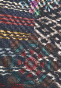 Tissu tissé complexe avec des motifs géométriques et floraux colorés en rouge, turquoise, jaune, blanc et marron sur un fond sombre.